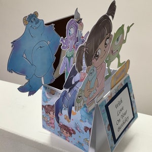 May include: A pop-up birthday card with a blue, purple, and green monster theme. The card features a blue monster, a purple monster, a green monster, and a girl with pigtails. The card is decorated with a blue and white pattern and the text 'With Love On Your Birthday'.