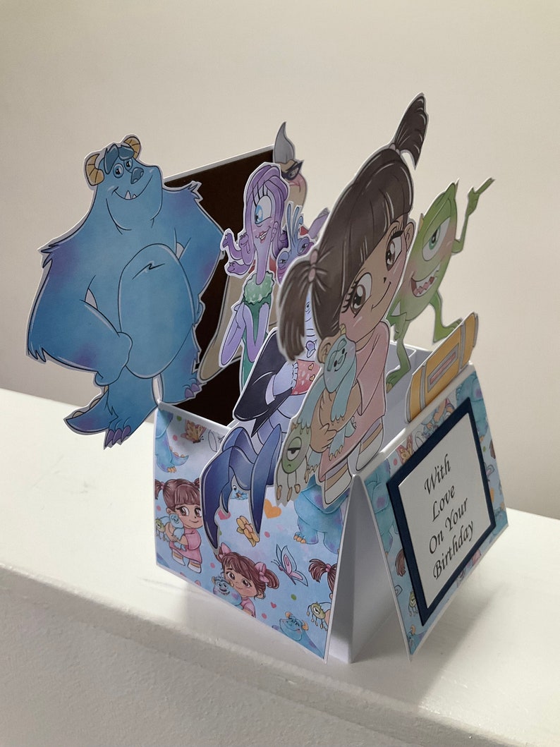 May include: A pop-up birthday card with a blue, purple, and green monster theme. The card features a blue monster, a purple monster, a green monster, and a girl with pigtails. The card is decorated with a blue and white pattern and the text 'With Love On Your Birthday'.