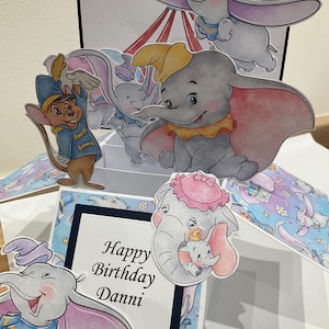 May include: A pop-up card featuring Dumbo the elephant and other characters from the Disney film. The card has a blue and white patterned background and includes the text "Happy Birthday Danni".