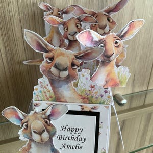 May include: A birthday card with a pop-up design, featuring a family of cartoon kangaroos. The card has the text "Happy Birthday Amelie". The kangaroos are depicted in shades of brown and tan, with large ears and expressive faces.