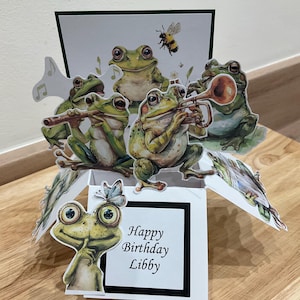 May include: A pop-up card featuring a 3D scene of frogs playing musical instruments. The card is open and the front reads "Happy Birthday Libby".