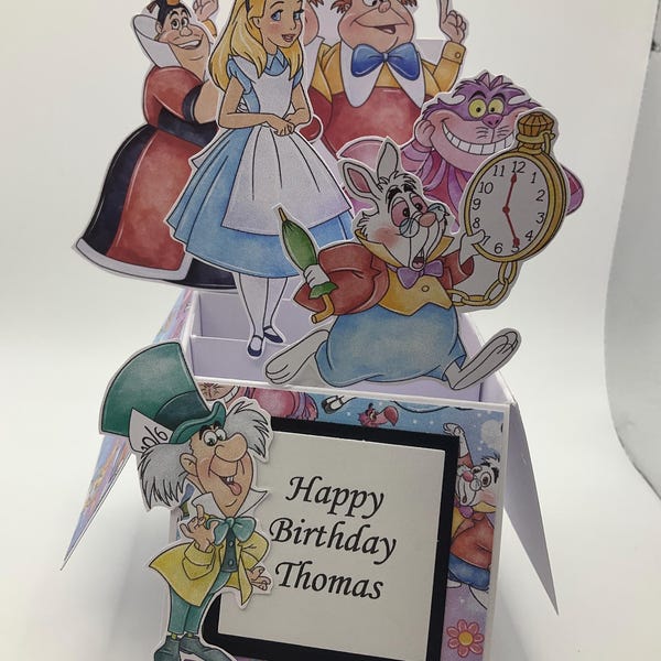 Peut inclure: Une carte de vœux pop-up 3D représentant des personnages d'Alice au pays des merveilles, dont Alice, la Reine de Cœur, le Lapin Blanc avec une montre à gousset, le Chat du Cheshire et le Chapelier Fou. Le texte Happy Birthday Thomas est inscrit.