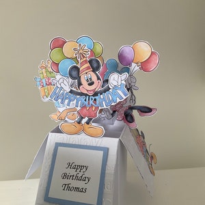 May include: A pop-up card featuring Mickey Mouse holding balloons and a banner that says "Happy Birthday". The card is open and has a blue and white patterned background with the text "Happy Birthday Thomas" on the front.