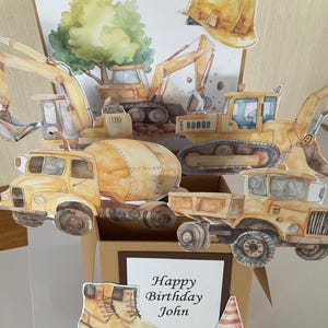 May include: A handmade birthday card with a pop-up construction theme. It features yellow construction vehicles, a hard hat, and a tree. The card includes the text "Happy Birthday John" on a white rectangle, a pair of work boots and a traffic cone.