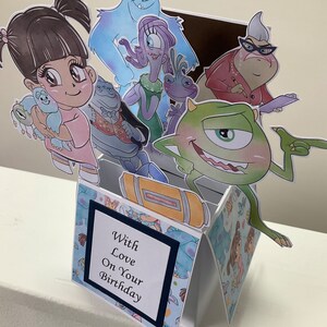 May include: A pop-up card with a blue, green, and white patterned background. The card features characters from the movie Monsters Inc. including Sully, Mike, Boo, Randall, and Celia. The card has a blue and white card with the text 'With Love On Your Birthday'.