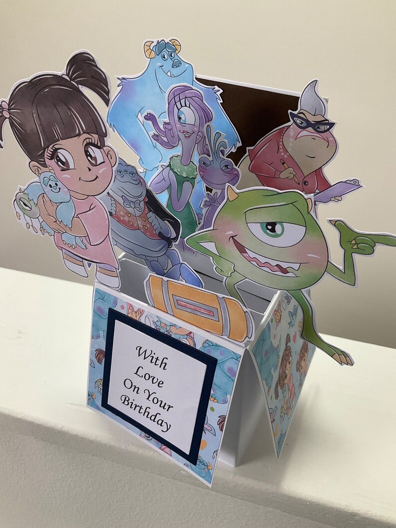 May include: A pop-up card with a blue, green, and white patterned background. The card features characters from the movie Monsters Inc. including Sully, Mike, Boo, Randall, and Celia. The card has a blue and white card with the text 'With Love On Your Birthday'.