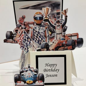 May include: A 3D pop-up greeting card featuring Formula 1 racing imagery. The card displays race cars, drivers in helmets, checkered flags, and a gold trophy. A square white panel on the front features the black printed text: Happy Birthday Jenson.