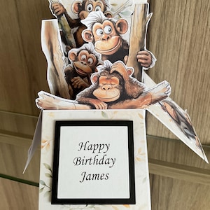 May include: A pop-up card with a jungle theme featuring four monkeys. The card is open and the monkeys are peeking out from behind a tree. The card has a black frame with the text "Happy Birthday James" inside.