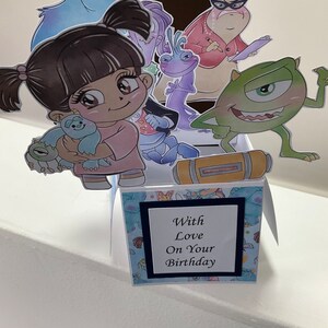 May include: A pop-up birthday card with a blue, green, and purple monster theme. The card features a pop-up of a girl holding a small blue monster. The card also features a pop-up of a green monster with one eye and a pop-up of a purple monster with a long neck. The card has a blue and white patterned background and a black frame with the text 'With Love On Your Birthday'.