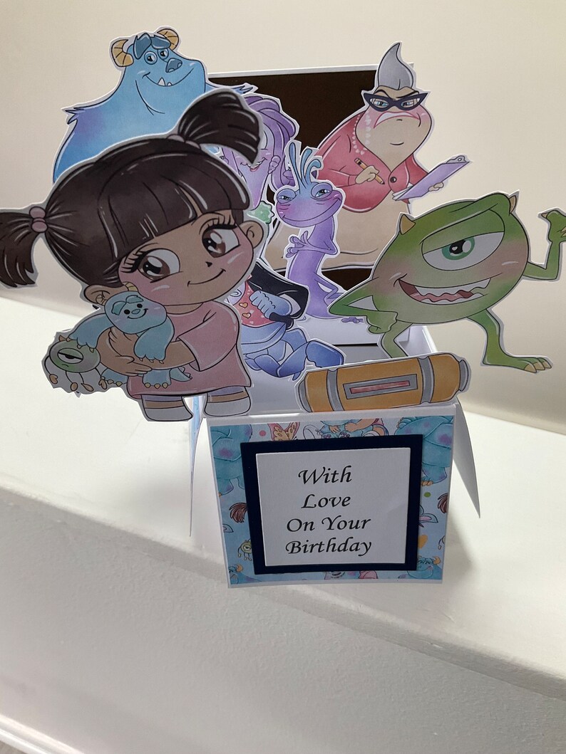 May include: A pop-up birthday card with a blue, green, and purple monster theme. The card features a pop-up of a girl holding a small blue monster. The card also features a pop-up of a green monster with one eye and a pop-up of a purple monster with a long neck. The card has a blue and white patterned background and a black frame with the text 'With Love On Your Birthday'.