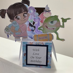 May include: A pop-up card with a blue and white floral pattern. The card features characters from the movie Monsters Inc. including Sully, Mike, Boo, and Randall. The card has a pop-up feature with the characters standing on a yellow box. The card has a white square with black trim and the text 'With Love On Your Birthday'.