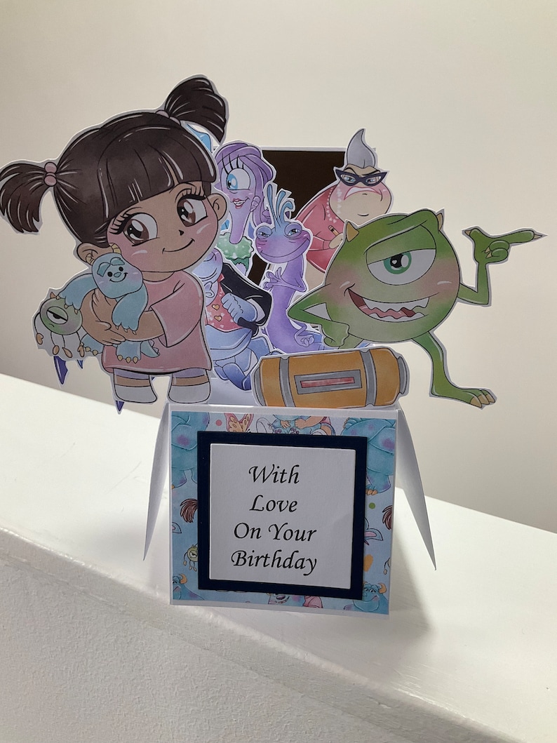May include: A pop-up card with a blue and white floral pattern. The card features characters from the movie Monsters Inc. including Sully, Mike, Boo, and Randall. The card has a pop-up feature with the characters standing on a yellow box. The card has a white square with black trim and the text 'With Love On Your Birthday'.