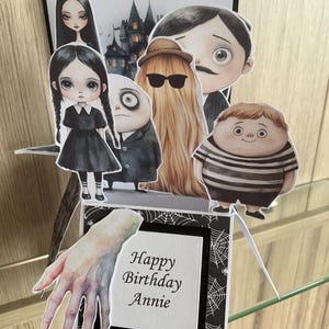 May include: A pop-up birthday card featuring illustrations of characters from The Addams Family. The card includes a black and white background with a hand illustration and the text "Happy Birthday Annie". The characters include Wednesday Addams, Gomez Addams, and Uncle Fester.