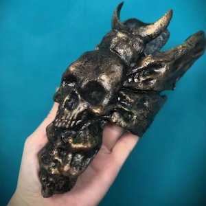 Puede incluir: Una escultura de calavera de color bronce con cuernos y elementos en forma de alas. La calavera tiene rasgos faciales detallados y se sostiene sobre un fondo turquesa. La escultura parece estar hecha de un material pesado.