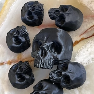 May include: A collection of seven black skull figurines. The skulls are detailed with visible teeth, eye sockets, and nasal cavities. The skulls are arranged on a light-colored surface with brown and yellow veining.