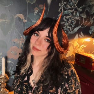 Puede incluir: Una persona con un par de cuernos grandes y curvados de color marrón rojizo. Los cuernos son el punto focal, sobre cabello oscuro. La persona lleva una camisa oscura con estampado floral. El fondo presenta un papel pintado floral.