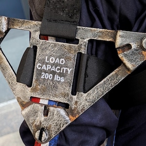 Puede incluir: Un componente de arnés de metal desgastado con el texto "LOAD CAPACITY 200 lbs." El componente está unido a una correa negra y presenta una franja roja, blanca y azul. El metal tiene un aspecto desgastado y envejecido.