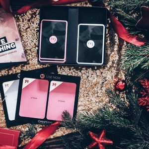 May include: A game called "Love Matching" in a black box with a red and black mask design. The box is on a gold sparkly surface with a red ribbon and a Christmas tree branch. There are two pink and white cards with the text "(V)" on them. There are also two pink cards with the text "USE NOW" and "2nd Date" on them.