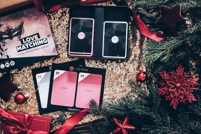 May include: A game called "Love Matching" in a black box with a red and black mask design. The box is on a gold sparkly surface with a red ribbon and a Christmas tree branch. There are two pink and white cards with the text "(V)" on them. There are also two pink cards with the text "USE NOW" and "2nd Date" on them.