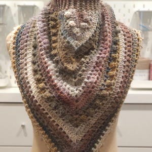 Peut inclure: Un châle crocheté à la main, multicolore, présenté sur un mannequin. Le châle présente un motif texturé avec des nuances de marron, de beige et des touches de bleu et de rose. La forme triangulaire et le point complexe sont mis en évidence.