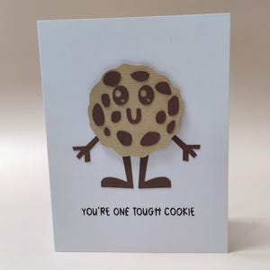 May include: A light blue card with a cartoon cookie illustration. The cookie is brown with dark brown spots and a smiling face. It has arms and legs. The text "YOU'RE ONE TOUGH COOKIE" is printed in black at the bottom of the card.