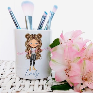May include: White ceramic makeup brush holder with a cartoon illustration of a girl with brown hair and blue eyes. The girl is wearing a white shirt, black pants, and brown boots. The text "Emily" is written below the illustration. The holder is filled with blue and pink makeup brushes.