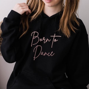 May include: Black hoodie with a front pocket and a drawstring hood. The words "Born to Dance" are written in a cursive, pink font across the chest. The model is wearing blue jeans.