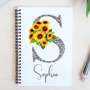 May include: A white spiral notebook with a silver glitter letter "S" decorated with yellow sunflowers and the name "Sophia" written in black cursive.
