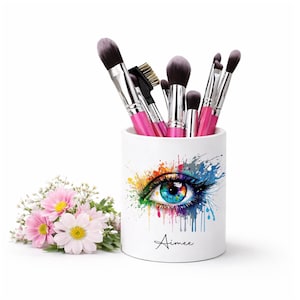 May include: A white ceramic makeup brush holder featuring a colorful eye design with a blue iris and rainbow accents. The holder contains various makeup brushes with pink handles. The name "Aimee" is written below the eye. A small bouquet of pink flowers is to the left.