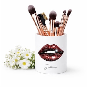 May include: White ceramic makeup brush holder featuring a red glitter lips design and the name "Jessica". The holder contains makeup brushes with rose gold handles and dark bristles. A small bouquet of white flowers is to the left.