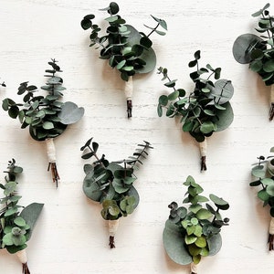 May include: Nine small bouquets of eucalyptus greenery tied with white twine, arranged on a white wooden surface.