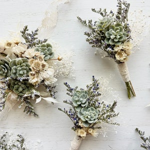 May include: Four small bouquets of dried flowers and succulents, tied with twine, on a white wooden background. The bouquets feature white flowers, green succulents, and sprigs of lavender.