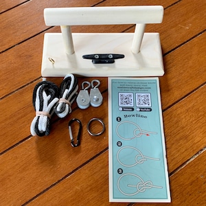 May include: A wooden knot tying station with a cleat and two vertical supports. Included are black and white ropes, pulleys, a carabiner, a metal ring, and a card with knot-tying instructions. The card has the text "Bowline".