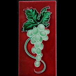 May include: A red ceramic tile with a green grape vine design. The grapes are green and white and the vine is green. The tile is a square shape.