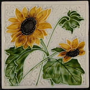 May include: Square ceramic tile with a sunflower design. The sunflowers have yellow petals and dark centres, with green leaves and stems. The background has a subtle floral pattern, creating a decorative and artistic piece.