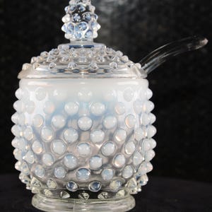 Puede incluir: Un tarro de vidrio transparente con tapa y cuchara. El tarro es blanco y tiene un patrón de hobnail. La tapa tiene un pomo en la parte superior. La cuchara está unida a la tapa. El tarro se asienta sobre una base de vidrio transparente.