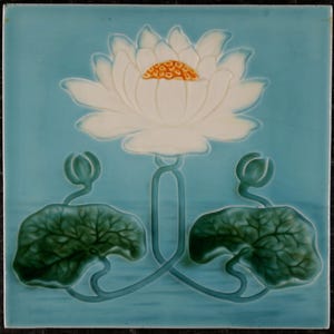 Op de afbeelding: Een vierkante keramische tegel met een reliëfontwerp van een witte lotusbloem met een geel hart. De bloem is gecentreerd boven twee donkergroene waterleliebladeren en twee gesloten knoppen, op een effen lichtblauwe achtergrond.