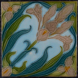 May include: A square ceramic tile featuring a central light brown iris flower with yellow accents, surrounded by dark green leaves and smaller floral buds. The background is a light blue colour with a visible crackle glaze finish throughout the surface.