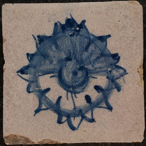 Puede incluir: Una baldosa cuadrada de color blanquecino con un diseño floral azul pintado a mano. El diseño presenta una espiral central rodeada de formas puntiagudas en forma de pétalos. La baldosa tiene bordes astillados, lo que sugiere edad y desgaste.