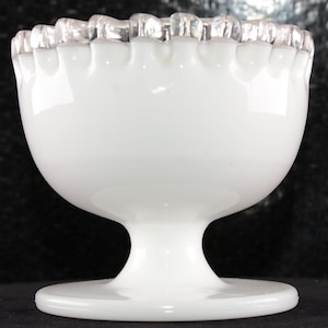 May include: A white, footed glass bowl with a ruffled rim. The bowl has a rounded body and a pedestal base. The rim is clear and has a scalloped edge. The bowl is likely vintage and is suitable for serving desserts or displaying small items.