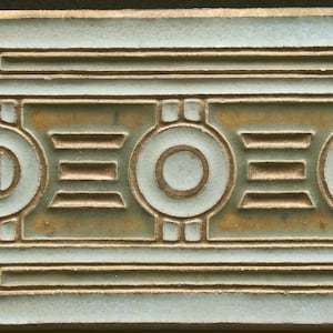 Op de afbeelding: Een keramische tegel met een geometrisch ontwerp in goud en wit op een lichtblauwe achtergrond. Het ontwerp bevat cirkels, vierkanten en lijnen.
