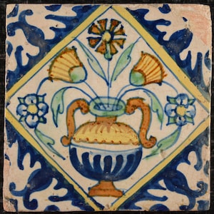 May include: A blue and white ceramic tile with a yellow and orange floral design. The tile features a vase with three flowers and a yellow and orange floral pattern in the corners.