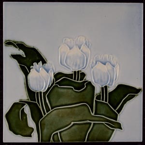 May include: Square ceramic tile featuring a raised relief design of three white tulip buds with green leaves. The background is a pale blue. The tile is framed with a dark border. The floral design is a classic decorative element.