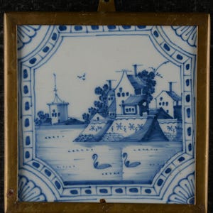 Può includere: Piastrella in ceramica blu e bianca con una scena di paesaggio, incorniciata in metallo color oro. La scena raffigura edifici, acqua e cigni. La piastrella ha un bordo decorativo con motivi blu.