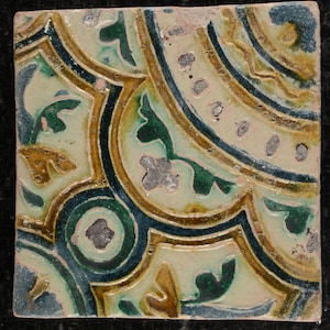 May include: A square ceramic tile featuring a repeating pattern of green, blue, and gold floral designs. The tile has a white background and a slightly textured surface.