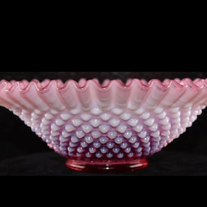 Puede incluir: Un cuenco de cristal rosa y blanco con borde ondulado. El cuenco tiene una base roja oscura y una superficie texturizada. El cuenco es probablemente vintage y es una pieza decorativa.