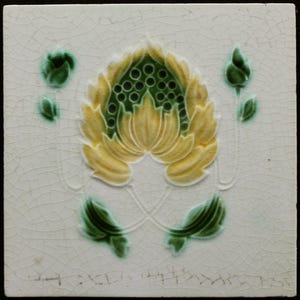 May include: A white ceramic tile with a green and yellow floral design. The design features a large stylized flower with a green center and yellow petals. The tile has a crackled glaze.