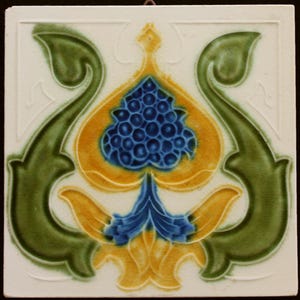 May include: A ceramic tile with a stylized floral design. The tile features a central motif of a blue and yellow flower with green leaves. The tile is decorated with a white background.
