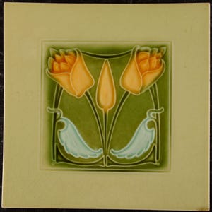 May include: Square ceramic tile with a floral design. The tile features two orange tulip-like flowers and a central bud against a green background. Light blue leaf-like shapes are at the bottom. The tile is framed by a light green border.