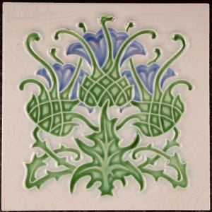 Op de afbeelding: Een vierkante keramische tegel met een witte achtergrond en een reliëfontwerp van een distelplant. De plant heeft groene stengels en bladeren met een gearceerde textuur en blauwe bloemhoofdjes. Het witte oppervlak heeft een craquelé glazuur.
